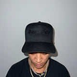 Person wearing a black cap with 'Good Boy' text and a black shirt with a logo on a plain background