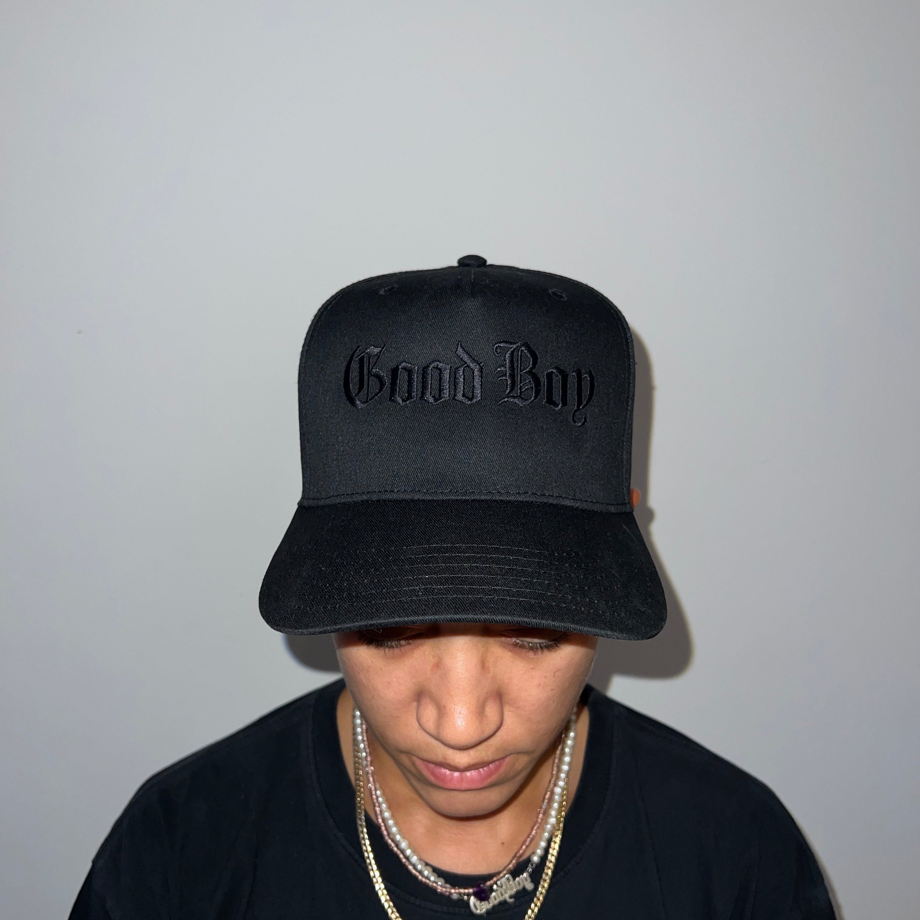 Person wearing a black cap with 'Good Boy' text and a black shirt with a logo on a plain background